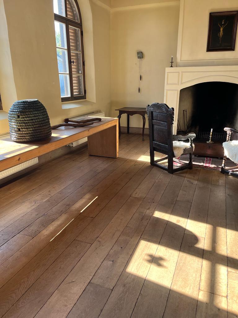 Schaduw van een absumo op houten vloer, met zicht op lange houten tafel, openhaard en houten stoel met hoge rugleuning en schapenvacht.